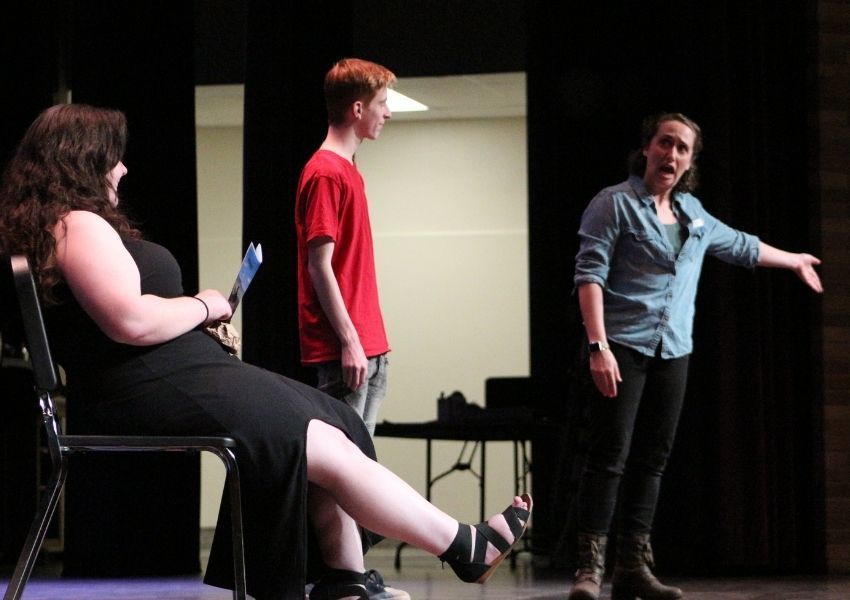 An actor gestures on stage to two students looking on. 