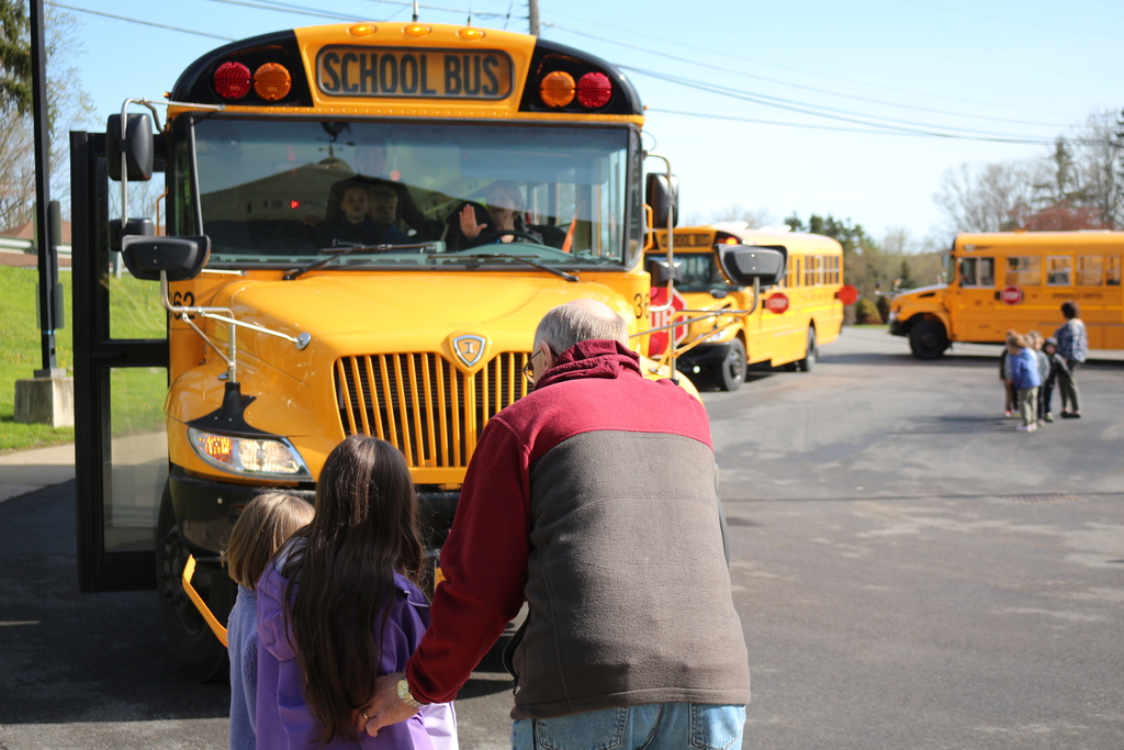 A bus driver gestures to a student that it is safe to cross. The student, with a bus monitor, looks up at the bus driver.